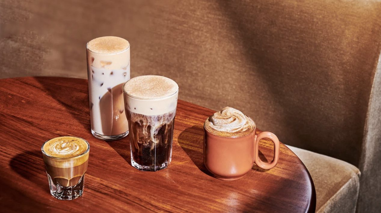 Two Starbucks autumn drinks, a Pumpkin Spice Latte and a Pecan Oatmilk Cortado, sitting side by side on a wooden coffee table.