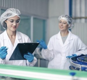 Two food safety specialists in white labcoats reviewing a checklist on a clipboard in a food production facility, representing ISO 22000 standard compliance.