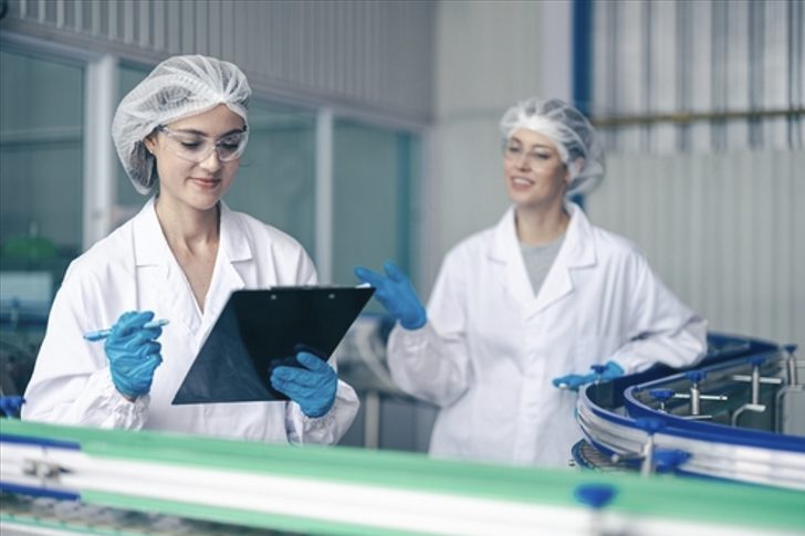 Two food safety specialists in white labcoats reviewing a checklist on a clipboard in a food production facility, representing ISO 22000 standard compliance.
